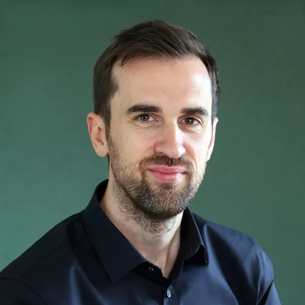 A man wearing a dark button-up shirt is positioned against a plain green background. The image focuses on the upper body and head, with the face blurred for privacy. The setting is minimalistic and professional, emphasizing simplicity and neutral tones.